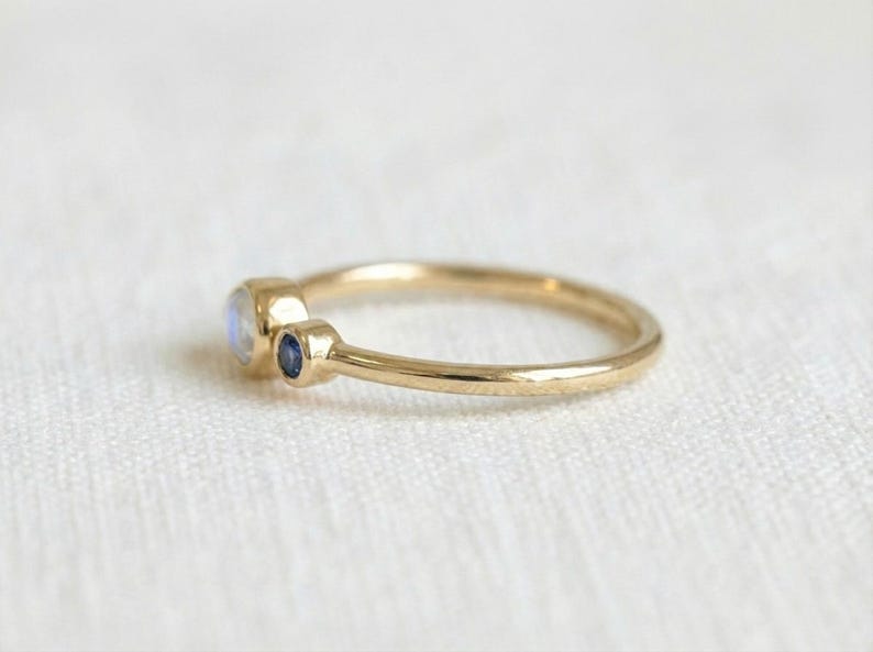 Pode incluir: Um delicado anel de ouro com uma pedra da lua redonda e iridescente e uma pequena safira azul escura. O anel tem uma banda fina e um design minimalista. As pedras preciosas s&atilde;o definidas em uma configura&ccedil;&atilde;o de moldura.