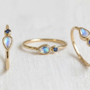 Pode incluir: Tr&ecirc;s an&eacute;is de ouro com engastes de pedras preciosas. Cada anel apresenta uma pedra da lua iridescente em forma de p&ecirc;ra combinada com uma pequena safira azul redonda. Os an&eacute;is s&atilde;o exibidos em uma superf&iacute;cie branca.