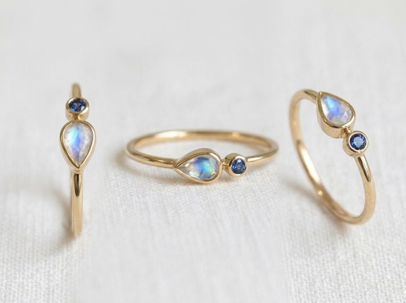 Pode incluir: Tr&ecirc;s an&eacute;is de ouro com engastes de pedras preciosas. Cada anel apresenta uma pedra da lua iridescente em forma de p&ecirc;ra combinada com uma pequena safira azul redonda. Os an&eacute;is s&atilde;o exibidos em uma superf&iacute;cie branca.
