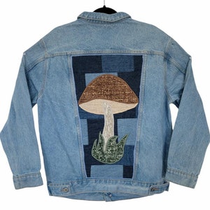Puede incluir: Una chaqueta vaquera azul claro con un parche de hongo en la espalda. El hongo está hecho de tela marrón y verde y está rodeado de un patchwork de mezclilla azul.