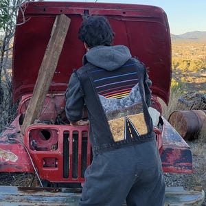 Op de afbeelding: Een persoon met een grijze hoodie en zwart vest met een kleurrijk ontwerp werkt aan een rode Jeep met de motorkap open. De Jeep staat buiten geparkeerd in de buurt van banden en bomen.