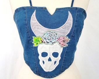 May include: A denim corset top with thin straps. The front features a white skull design with a gray crescent moon above it. Colorful faux succulents are placed above the skull. The top is displayed on a mannequin.