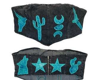 May include: A black denim corset with a zipper closure and turquoise blue embroidered designs. The designs include a wolf, stars, cacti, and crescent moons.