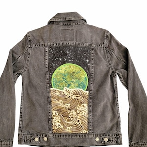 Op de afbeelding: Grijze spijkerjas met een geschilderd ontwerp op de rug. Het ontwerp toont een groen-gele planeet, een sterrenhemel en gestileerde golven in bruin- en goudtinten. De jas heeft een klassieke kraag, knopen en zakken.