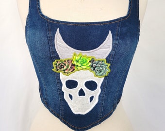 May include: A blue denim corset with a white skull and succulent flower crown embroidered on the front.