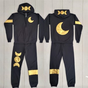 May include: Black hooded sweatshirt and sweatpants set with gold moon and triple moon phases design. The sweatshirt has a zipper closure and the sweatpants have gold stripes on the legs.