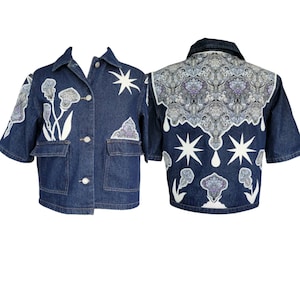 Peut inclure: Veste en jean bleu foncé à manches courtes et fermeture boutonnée. La veste présente des motifs floraux et étoilés blancs, ainsi qu'un panneau décoratif à motifs au dos. Deux poches avant ajoutent de la fonctionnalité.