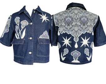 May include: A dark blue denim jacket with short sleeves and a button-up front. The jacket features white floral and star designs, along with a decorative patterned panel on the back. Two front pockets add functionality to the design.