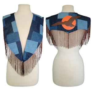 May include: A denim vest with a patchwork design in shades of blue. The vest features brown fringe along the bottom and a leather applique of a bird in front of an orange sun on the back. The vest is displayed on a mannequin.