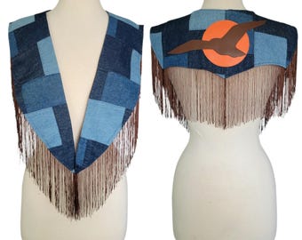 May include: A denim vest with a patchwork design in shades of blue. The vest features brown fringe along the bottom and a leather applique of a bird in front of an orange sun on the back. The vest is displayed on a mannequin.