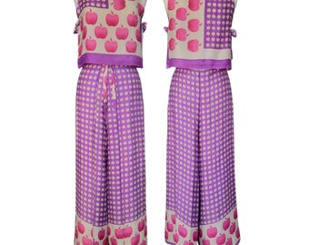 May include: A two-piece outfit featuring a sleeveless top and wide-leg pants. The top and pants are primarily purple with a white polka dot pattern. The top has a square panel with pink apple graphics. The pants have a matching apple border.
