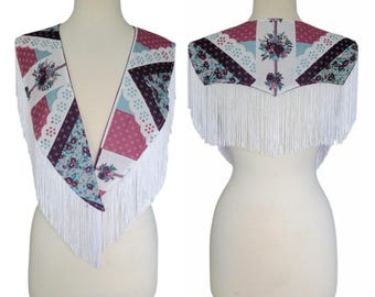 Peut inclure: Un châle présentant un motif patchwork avec des imprimés floraux et géométriques dans des tons bordeaux, bleu clair et blanc. Le vêtement possède une encolure en V profonde et est orné de longues franges blanches sur les bords, sur mannequin.