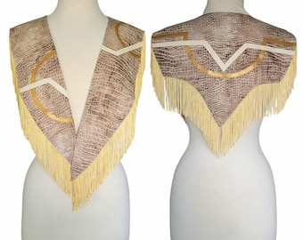May include: A brown and tan faux snakeskin print capelet featuring a V-neckline, cream-colored fringe trim along the edges, and decorative gold and white geometric piping accents. The garment is displayed on a mannequin from both front and back views.