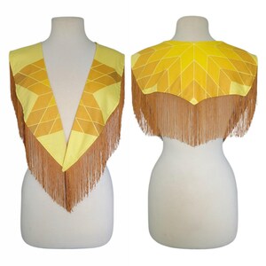 May include: A yellow and gold geometric patterned capelet featuring a deep V-neckline and long, flowing tan fringe trim along the edges. The garment displays a star-like design on the back and front panels, showcasing a vibrant color palette.