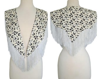 May include: A triangular shawl featuring a cream, black, and gray geometric pattern with long white fringe trim along the edges. The garment is displayed on a mannequin, showing both the front view with open panels and the back view with a pointed hem.