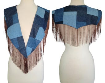 May include: A denim vest with a patchwork design in shades of blue. The vest features long brown fringe along the bottom edge. The vest is displayed on a mannequin.
