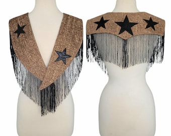 May include: A brown and black fringed shawl with three black stars on the back and two on the front. The shawl is displayed on a mannequin. The fringe is black and hangs down from the bottom of the shawl.