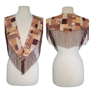 May include: A V-neck shawl featuring a patchwork design of various brown, tan, and cream geometric patterns. The garment is adorned with long, thin brown fringe along the edges, creating a layered look that drapes over the shoulders and chest.