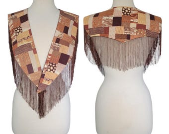 Peut inclure: Un châle à col en V présentant un motif patchwork composé de divers motifs géométriques marron, beige et crème. Le vêtement est orné de longues franges marron fines sur les bords, créant un aspect superposé qui tombe sur les épaules.