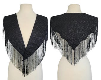 May include: A black, triangular shawl with a floral pattern and long, white fringe. The shawl is displayed on a mannequin, showcasing the front and back views of the garment. The fringe hangs from the bottom edge.