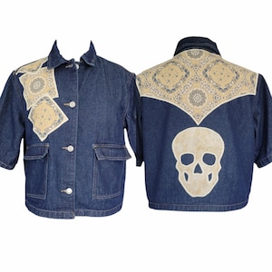 Könnte beinhalten: Dunkelblaue Jeansjacke mit kurzen Ärmeln, mit beigefarbenen Paisley-Patches auf den Schultern und dem Rücken. Der Rücken hat eine große Totenkopfapplikation. Die Jacke hat zwei Vordertaschen und drei Knöpfe.