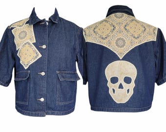 May include: Dark blue denim jacket with short sleeves, featuring beige paisley patches on the shoulders and back. The back has a large skull applique. The jacket has two front pockets and three buttons.