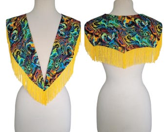 May include: A colorful, patterned shawl with a V-neck design and yellow fringe. The fabric features a swirling mix of blues, greens, oranges, and purples. The shawl is displayed on a white mannequin.