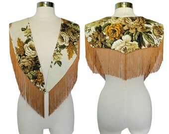 Peut inclure: Une cape d'épaules en tissu crème ornée d'un motif floral de roses et de feuillages jaunes, bruns et verts. Le vêtement est agrémenté de longues franges brun clair sur les bords inférieurs, visibles à la fois de face et de dos.