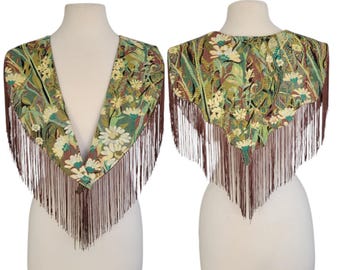 May include: A floral tapestry shawl with a V-neck design and brown fringe. The shawl features a pattern of white and yellow flowers with green and brown leaves. The fringe is long and hangs down from the bottom edge.