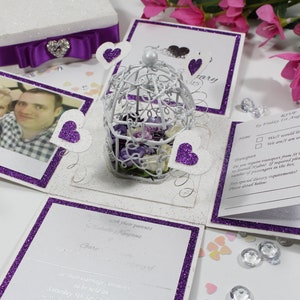 May include: A white and purple glittery box with a birdcage and hearts. The box is open and has a photo of a couple inside. The box is decorated with purple glitter and has a white ribbon with a purple bow. The text on the box says "RSVP by Friday 1st August 2017".