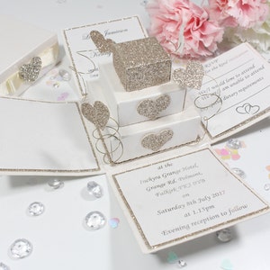 May include: A white and gold wedding invitation box with a pop-up design. The box is decorated with glitter and hearts. The invitation reads "at the Inchyra Grange Hotel, Grange Rd, Polmont, Falkirk PK2 0908 on Saturday 8th July 2017 at 1.15pm Evening reception to follow".