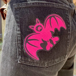 Peut inclure: Un jean noir en denim avec un patch de chauve-souris rose sur la poche arrière.