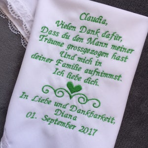 May include: White handkerchief with green embroidered text. The text includes a name, a thank you message, and a date. The handkerchief has a delicate lace trim along one edge. The overall design is simple and elegant.