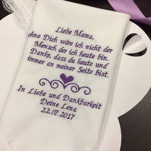 May include: White handkerchief with purple embroidered text that reads "Liebe Mama, ohne Dich wäre ich nicht der Mensch der ich heute bin. Danke, dass du heute und immer an meiner Seite bist. In Liebe und Dankbarkeit Deine Lena 22.07.2017"