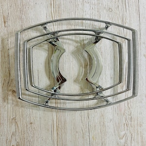 May include: A chrome-plated metal trivet with a rectangular frame and curved, decorative supports. The trivet has a spring-like design on the sides and is designed to protect surfaces from hot dishes. The trivet is shown on a light wood surface.