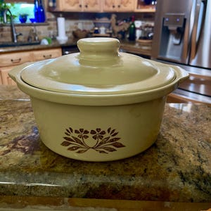 May include: A beige ceramic casserole dish with a lid. The dish has a brown floral design on the front. The casserole dish is sitting on a granite countertop.
