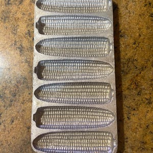 May include: A cast iron cornbread pan in the shape of six ears of corn. The pan is silver in color and has a loop at the top for hanging. The pan is designed to bake cornbread in the shape of corn on the cob.