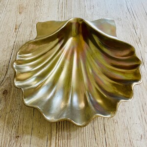 May include: A metallic, seashell-shaped decorative dish with a scalloped edge. The dish has a lustrous, iridescent finish with hints of gold and green. The dish is made of metal and sits on a light-colored wooden surface.