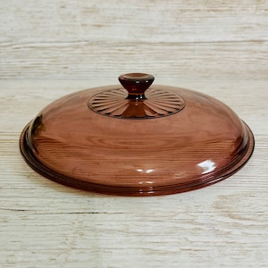 May include: A vintage, oval-shaped, glass lid in a muted purple hue. The lid features a central, circular design with radiating lines and a small, rounded knob. The glass has a slightly domed shape and a dark rim.