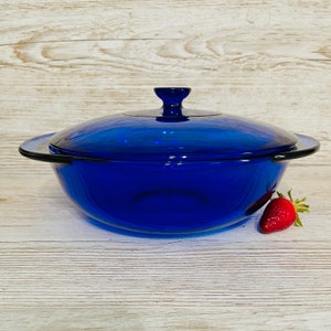 May include: A cobalt blue glass casserole dish with a lid. The dish has a round shape with a handle on each side. A red strawberry is next to the dish. The dish is on a light wood surface.