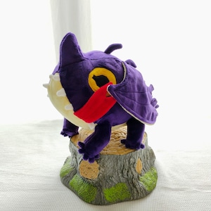 May include: Purple plush dragon with a yellow eye and red tongue, sitting on a gray tree stump with brown and green accents.