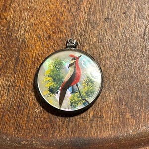 May include: Round pendant with a painted bird design. The bird has red, black, and white feathers, perched on a branch against a backdrop of green foliage. The pendant has a silver-colored metal frame and a bail for a chain.
