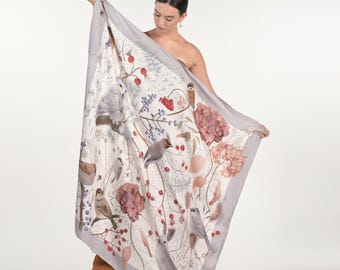 May include: A large, rectangular scarf with a nature-inspired design. The scarf features illustrations of birds, berries, and flowers in muted tones of gray, pink, and brown against a white background. The scarf is held up, showcasing its full design.