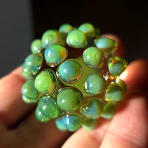 May include: A spherical glass bead with a textured surface, featuring numerous rounded, bubble-like protrusions. The bead is primarily green with hints of blue and yellow, and is held in a hand.