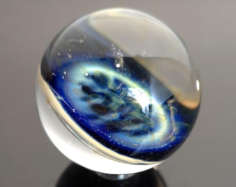 May include: A clear glass marble with an internal design of swirling blues, greens, and white, resembling a galaxy. The marble is spherical and sits on a reflective surface, casting a dark shadow.