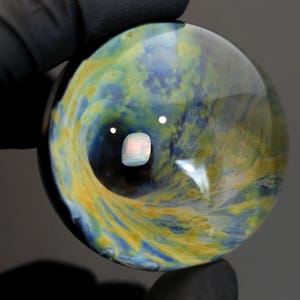 May include: A clear glass orb with swirling patterns of blue, yellow, and green. The orb has a reflective surface with a central, light-colored, square-shaped reflection. The orb is held by a gloved hand against a dark background.