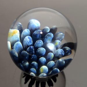 May include: A clear glass paperweight with a cluster of blue, oval-shaped inclusions. The inclusions have a marbled appearance with hints of yellow and white. The paperweight is spherical and reflects the light, creating a mirrored effect.