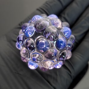 May include: A spherical glass art piece with a textured surface, featuring a pattern of rounded, bubble-like shapes. The colors range from deep blues and purples to lighter, translucent tones. The piece is held in a black gloved hand.