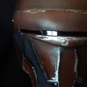 Può includere: Un elmetto marrone con un aspetto consumato e rovinato. L'elmetto ha una visiera con un bordo argentato e una sezione blu scuro nella parte inferiore dell'elmetto.