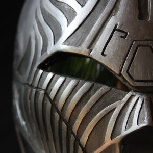 Könnte beinhalten: Nahaufnahme eines silbernen Helms mit einem strukturierten, gestreiften Design. Der Helm hat eine dunkle, rechteckige Augenöffnung.